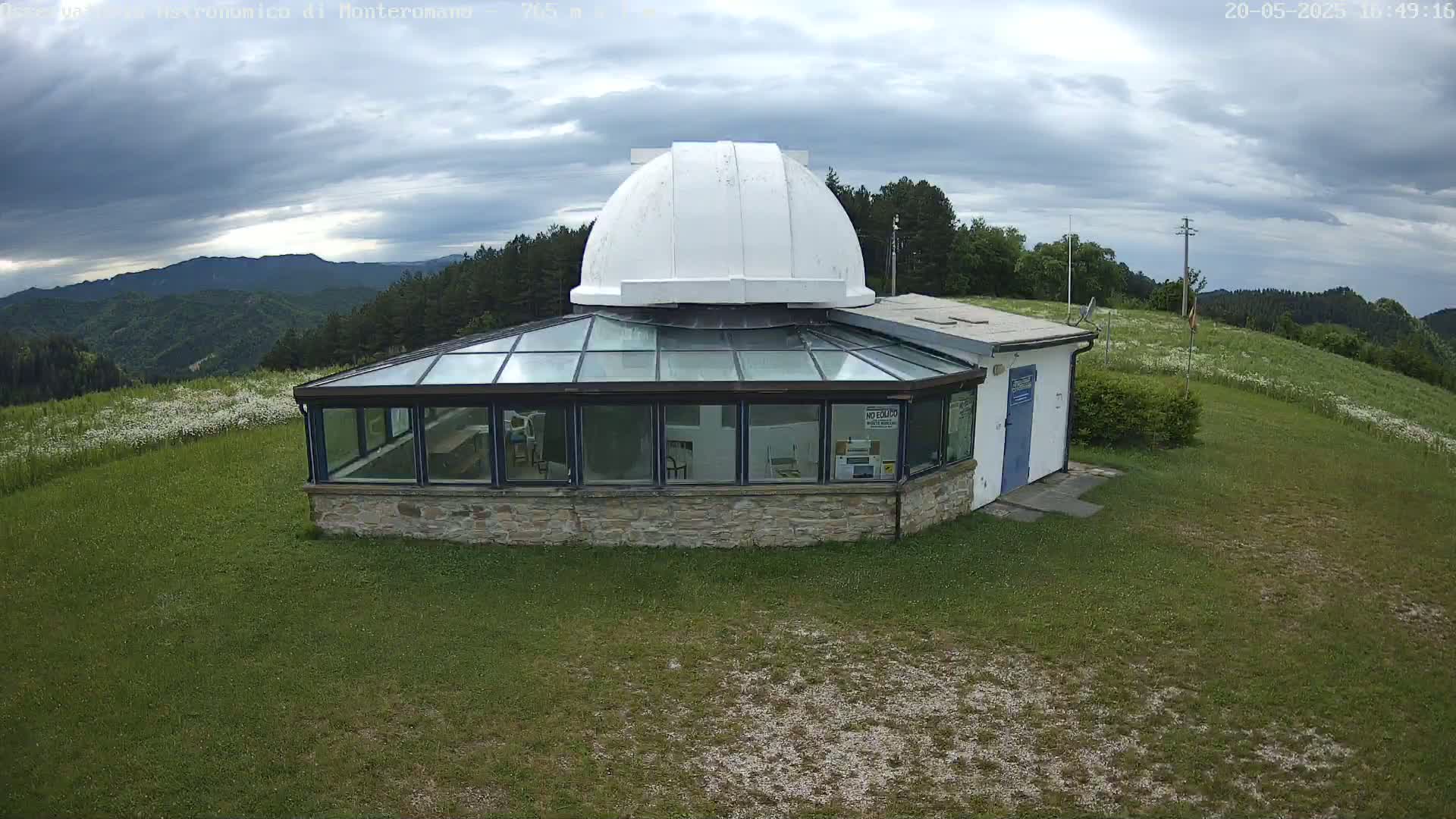A stone building with a glass roof and a white dome on top sits on a grassy hill overlooking a mountainous landscape under a partly cloudy sky.