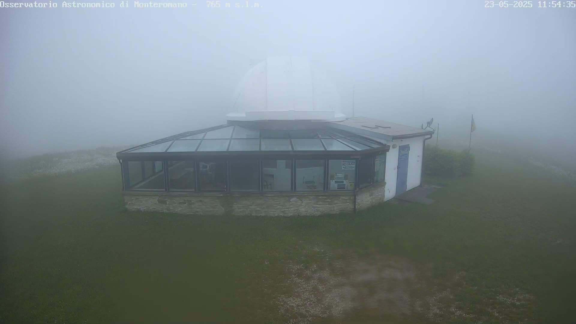 A white-domed observatory building with a glass-roofed addition sits on a grassy area shrouded in thick fog.