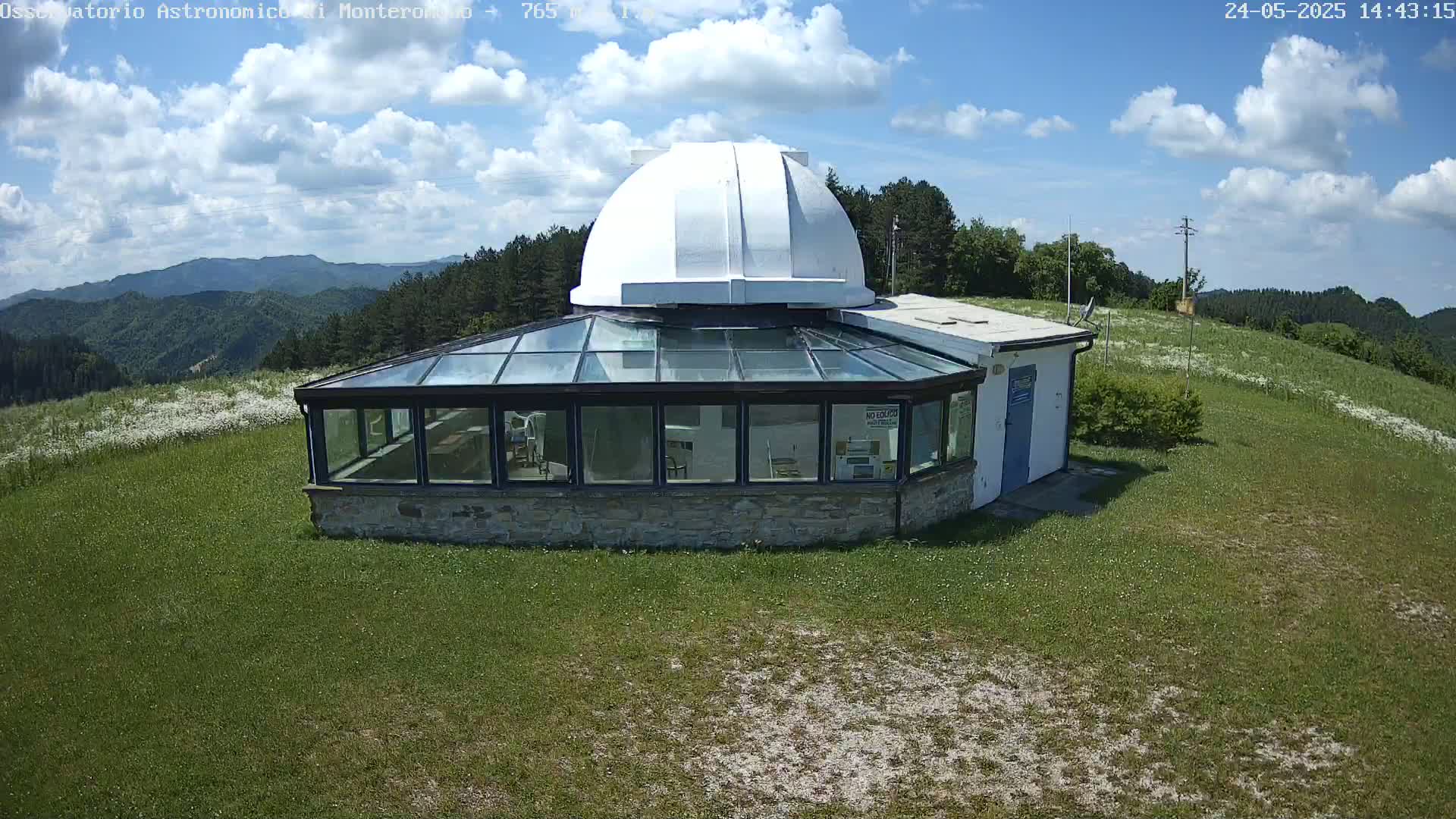A white domed observatory with a glass-roofed addition sits on a grassy hill overlooking a mountainous landscape under a partly cloudy sky.