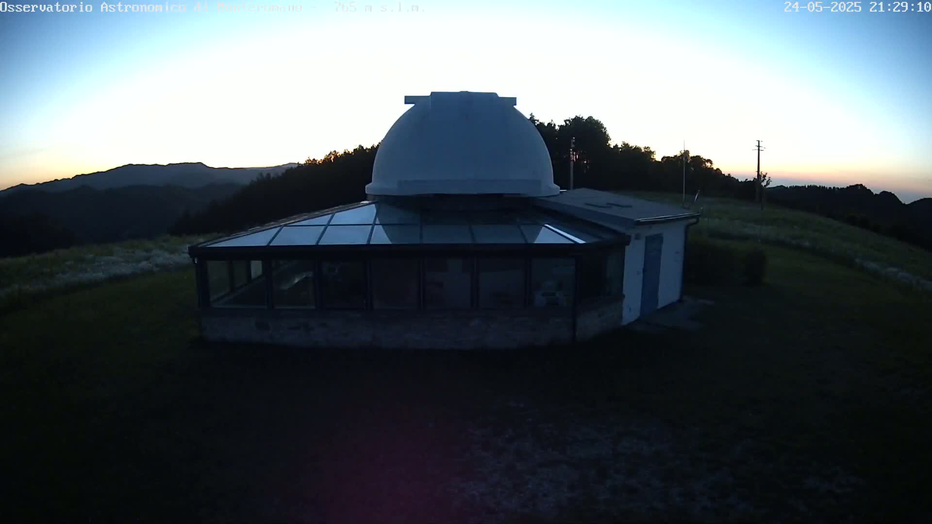 A white observatory dome sits atop a building with a glass-roofed addition, overlooking a landscape bathed in the twilight of a setting sun.