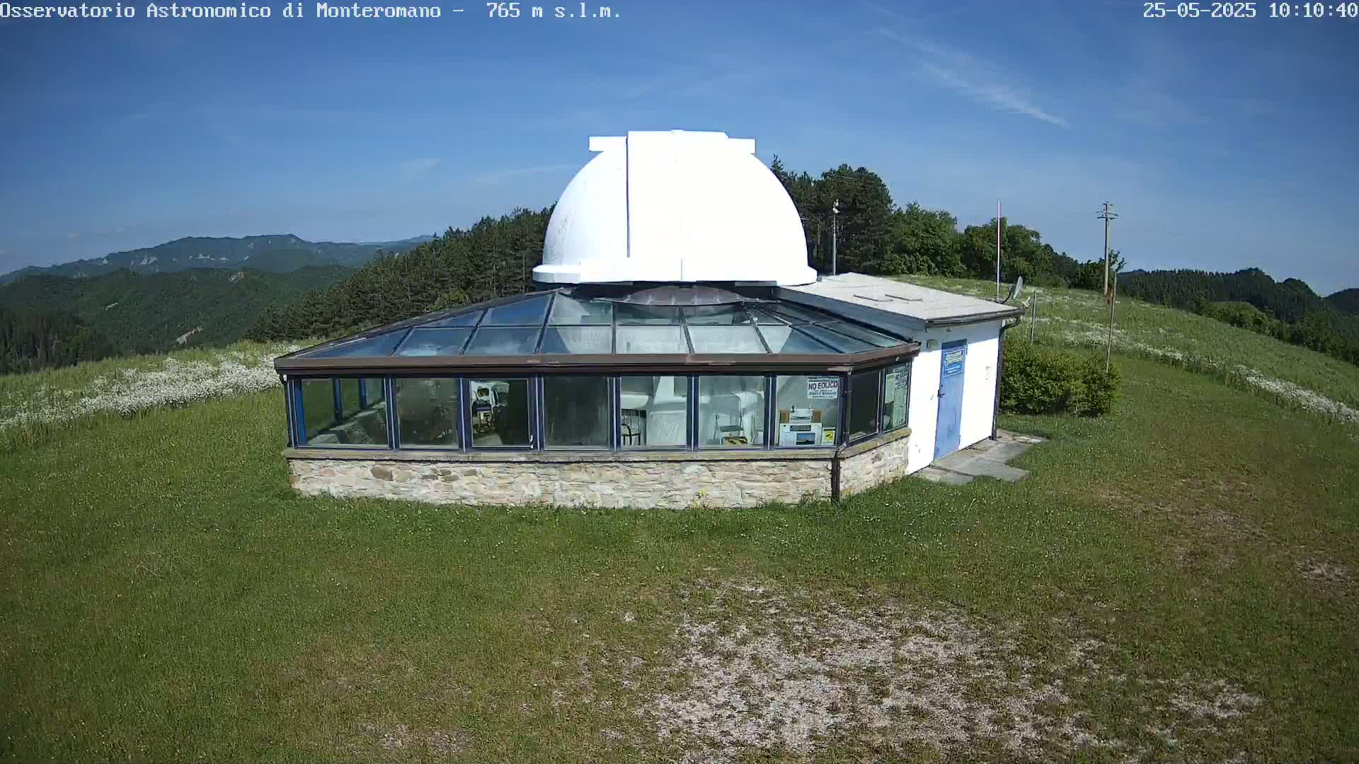 A white dome observatory sits atop a grassy hill under a clear blue sky, overlooking a mountainous landscape.