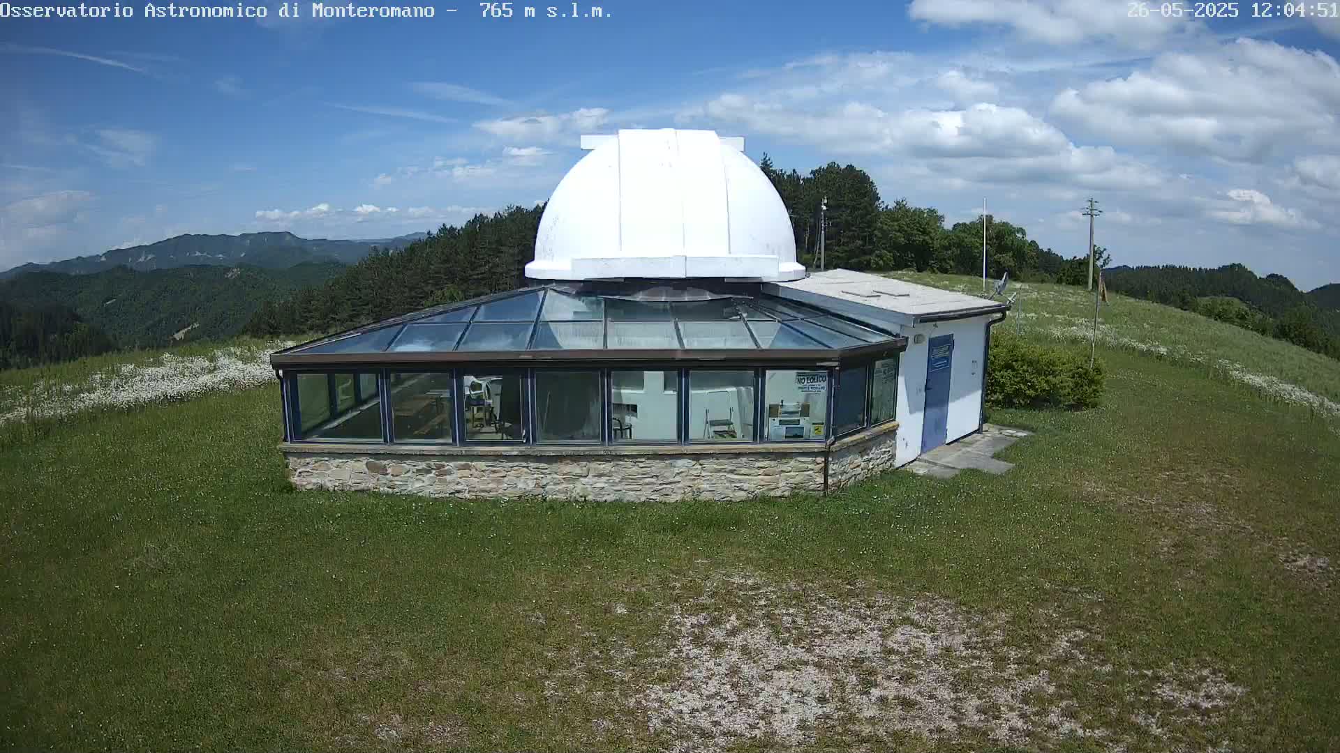 A stone building with a glass sunroom and a white dome sits atop a grassy hill on a partly sunny day, overlooking a mountain range in the distance.