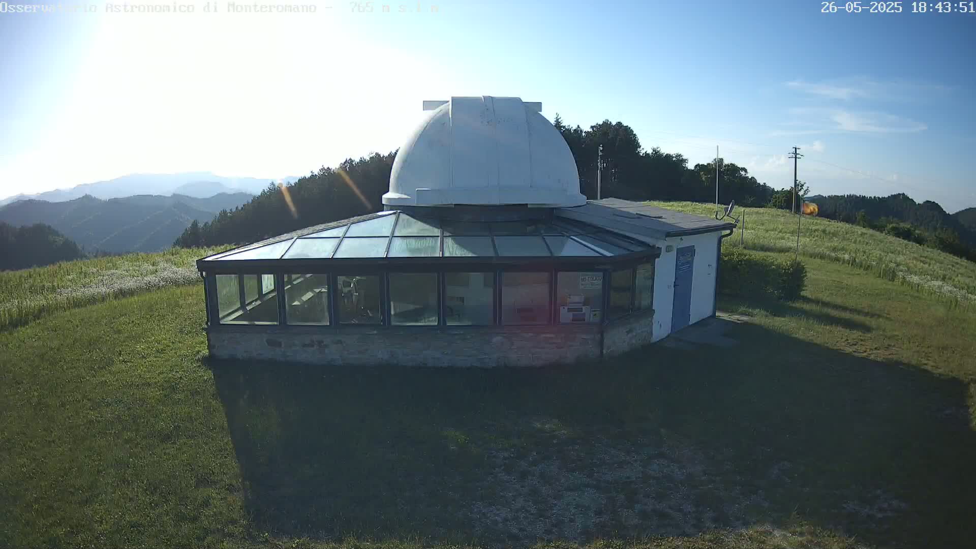 A white-domed observatory building with a glass-roofed addition sits on a grassy hill overlooking a mountainous landscape under a mostly clear sky.