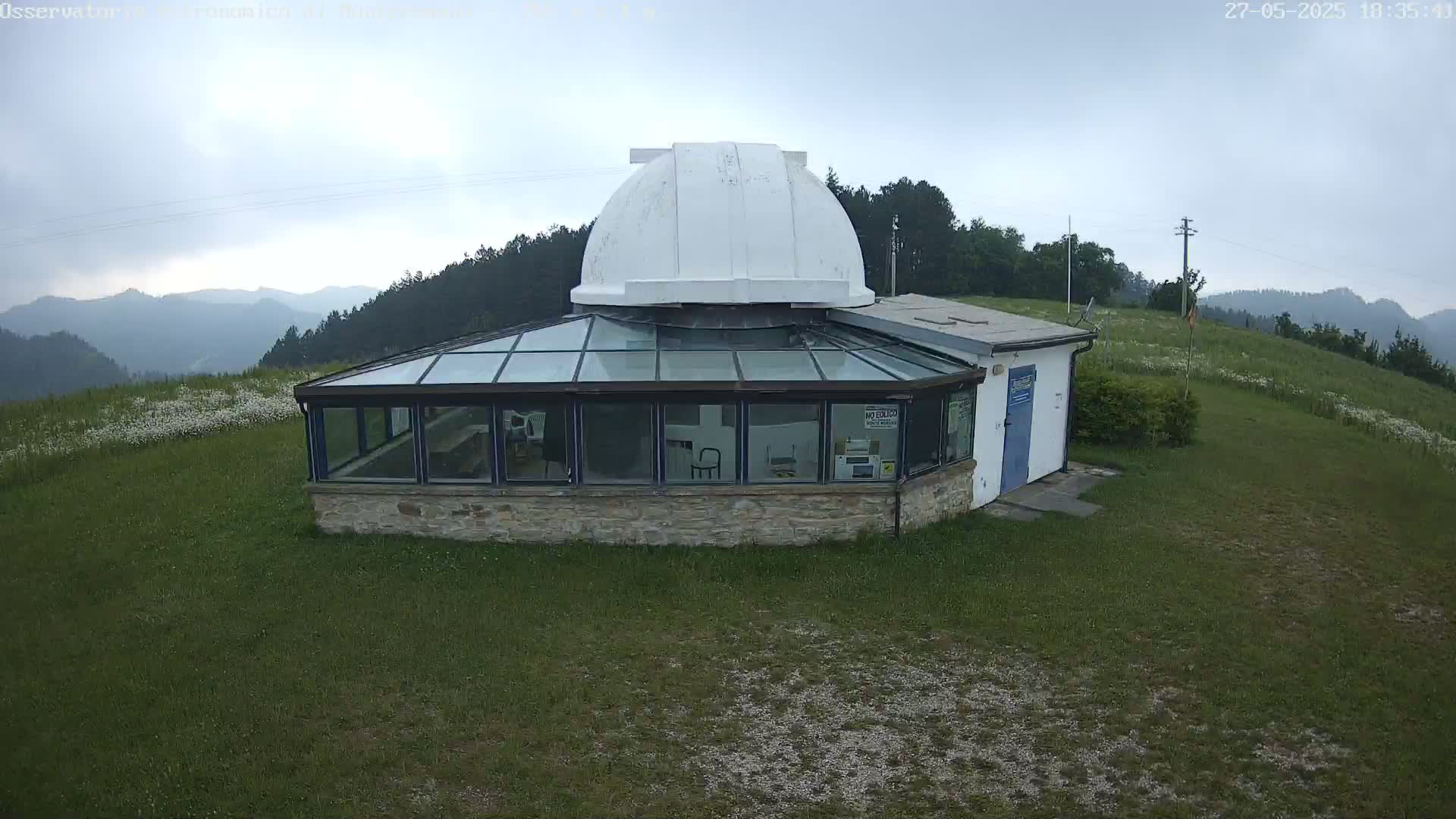 A white domed observatory building with a glass sunroom sits on a grassy hill overlooking a hazy mountain range under an overcast sky.