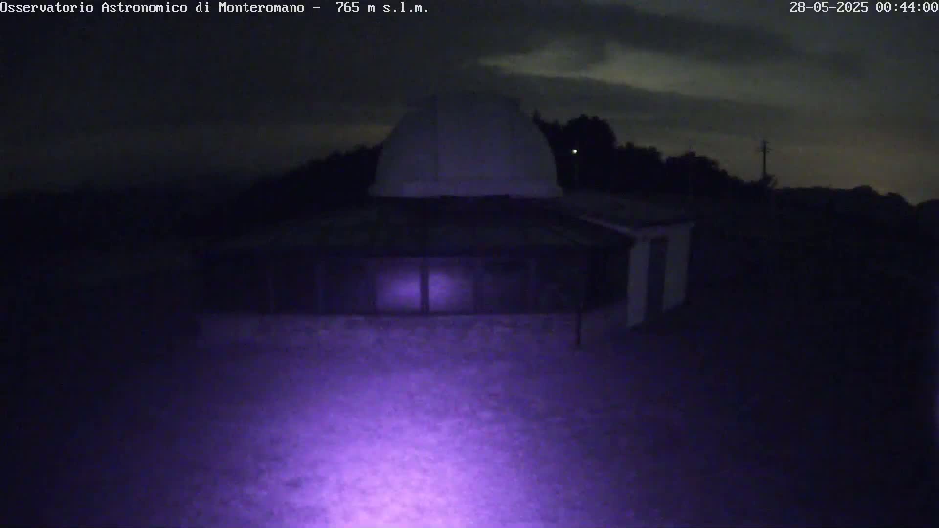 A low-light nighttime image shows a dark building with a dome-shaped roof, illuminated by a purple light source in the foreground, under a cloudy sky.