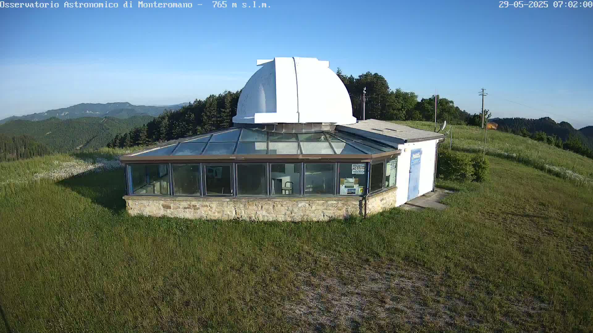 A white domed observatory building with a glass-walled addition sits on a grassy hilltop under a clear blue sky.