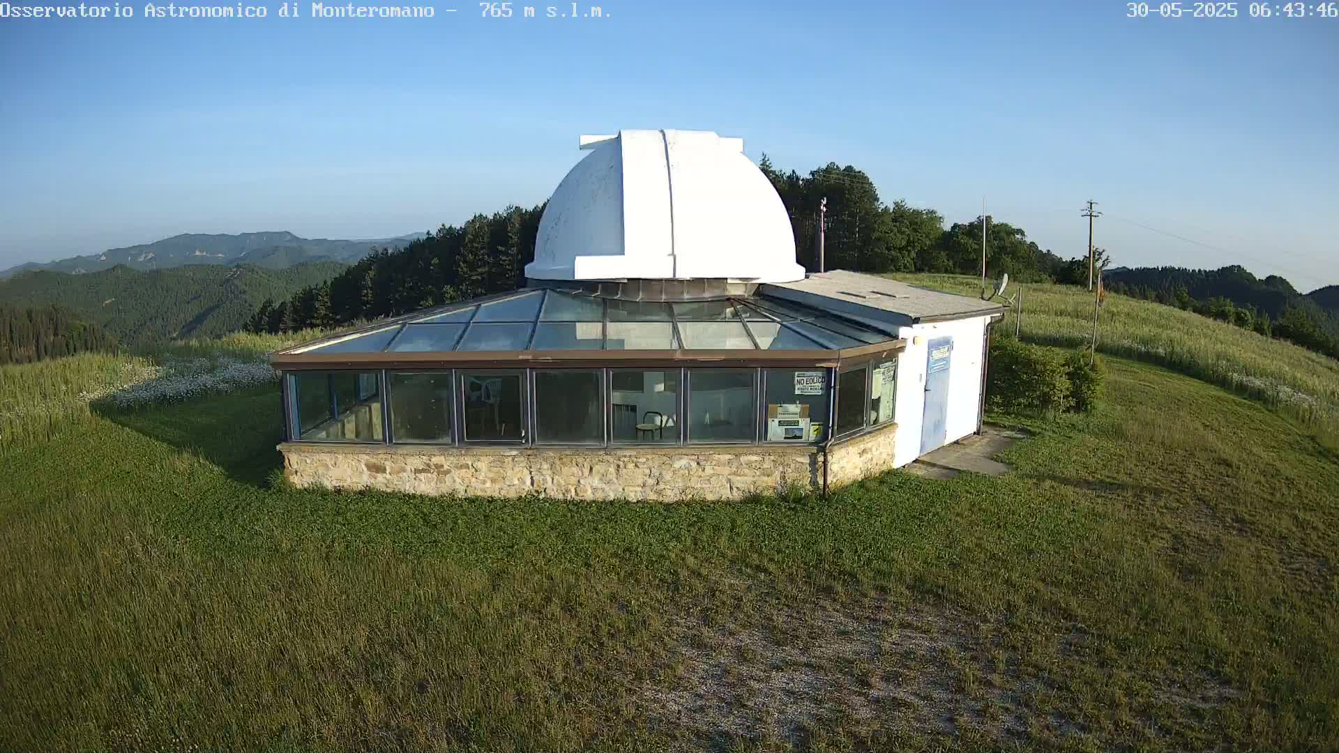 A white-domed observatory building with a glass-roofed addition sits on a grassy hilltop under a clear blue sky.