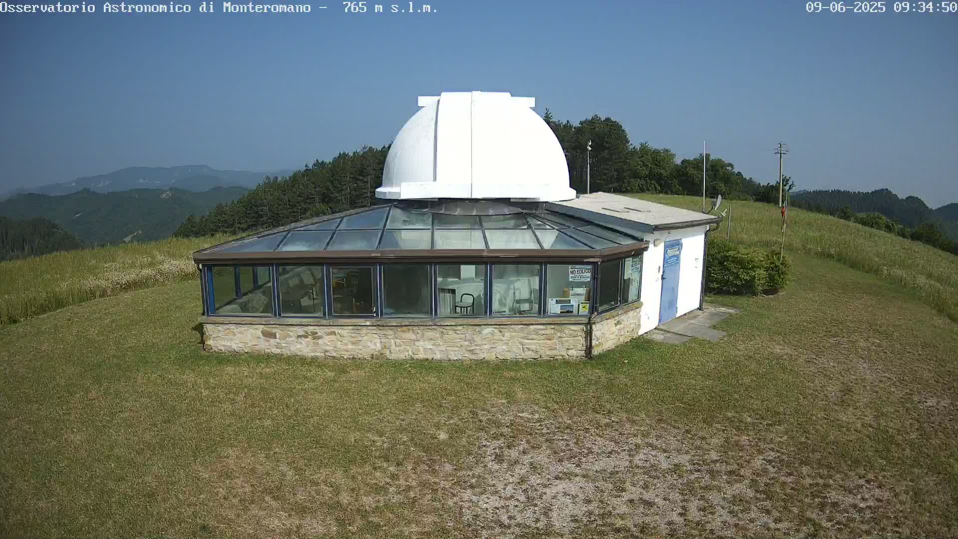 A stone building with a glass roof and a white dome on top sits on a grassy hill under a clear blue sky.
