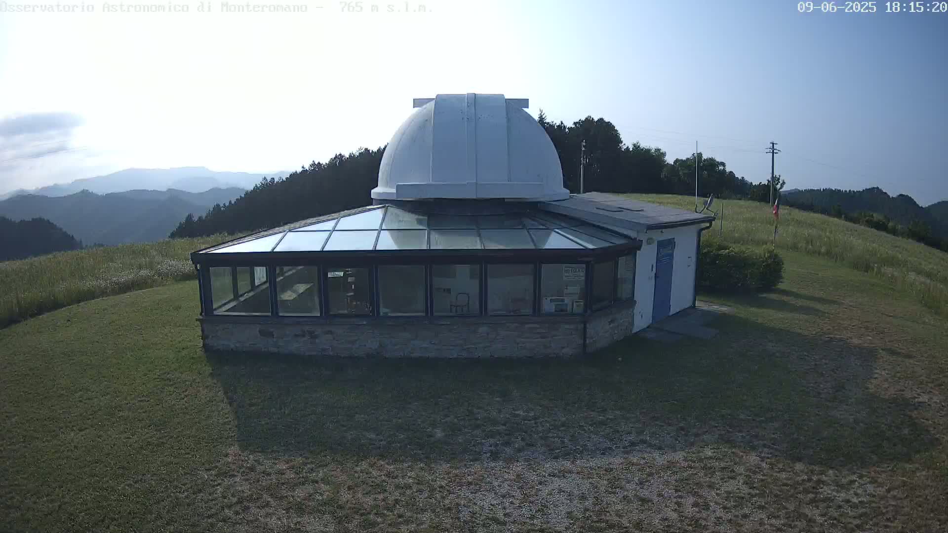 A white-domed observatory building with a glass-roofed addition sits on a grassy hill overlooking a hazy mountain range under a mostly clear sky.