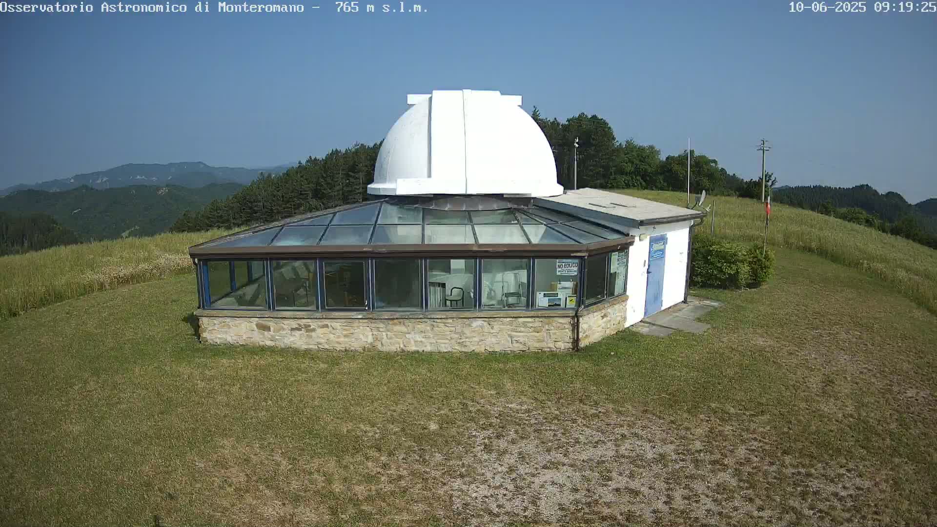 A stone building with a glass enclosure and a white dome sits on a grassy hilltop under a clear blue sky.