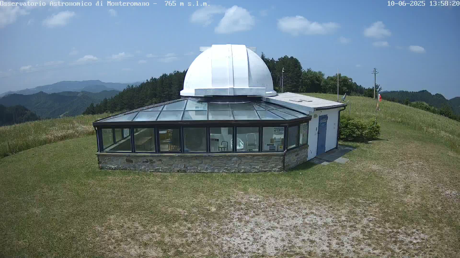A white dome observatory sits atop a stone building with a glass sunroom, overlooking a grassy hillside under a partly cloudy sky.