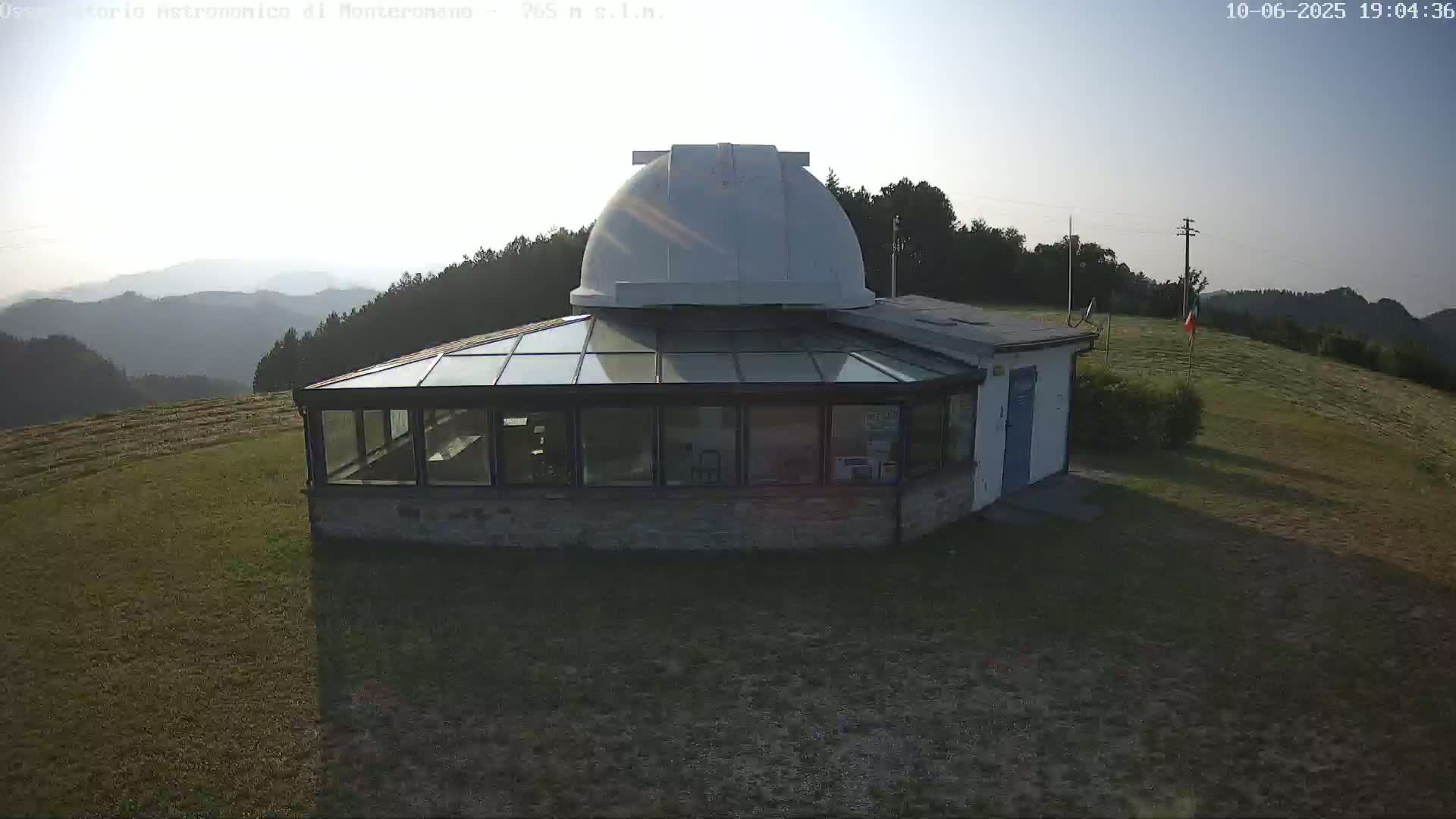 A stone building with a glass sunroom and a white dome on top sits on a grassy hill overlooking a hazy mountain range under a clear, bright sky.