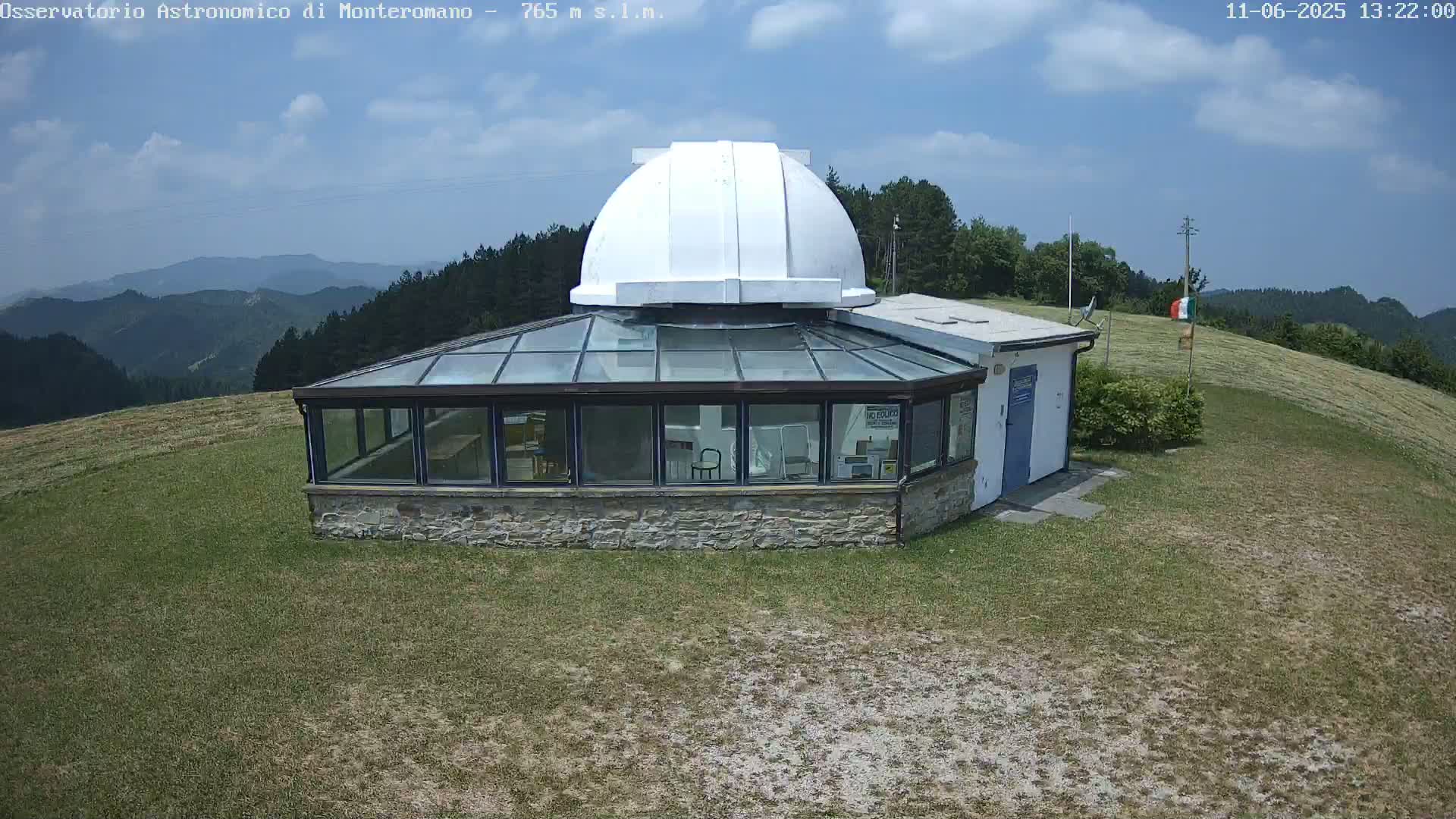A stone building with a glass roof and a white dome sits on a grassy hilltop under a partly cloudy sky, overlooking a range of distant hills.
