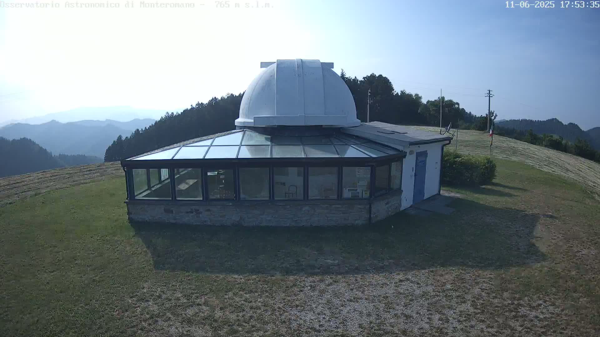 A white-domed observatory building with a glass-roofed addition sits on a grassy hill overlooking a hazy mountain range under a clear, sunny sky.