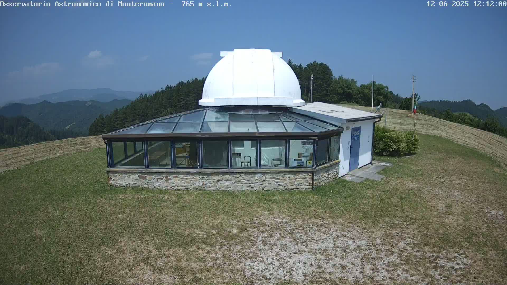 A white domed observatory building with a glass-roofed addition sits on a grassy hilltop under a clear blue sky.