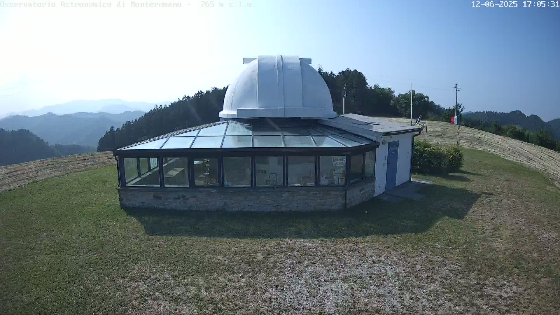 A white-domed observatory building with a glass sunroom is situated on a grassy hill overlooking a mountain range under a clear, sunny sky.