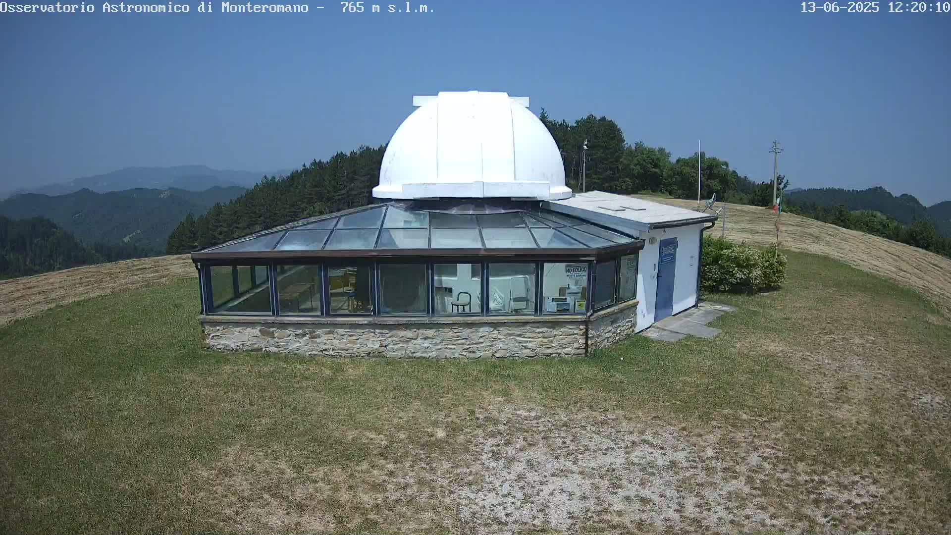 A stone building with a glass sunroom and a white dome sits atop a grassy hill on a clear, sunny day, overlooking a mountainous landscape.