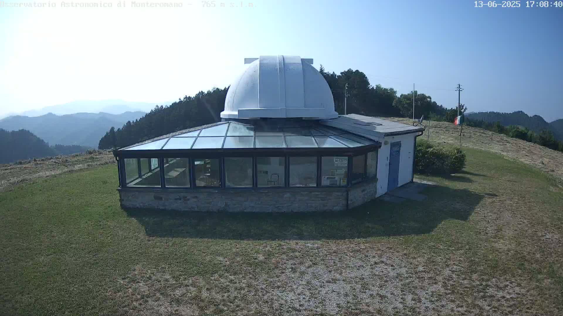 An observatory with a white dome sits atop a grassy hill on a sunny day, overlooking a distant mountain range.