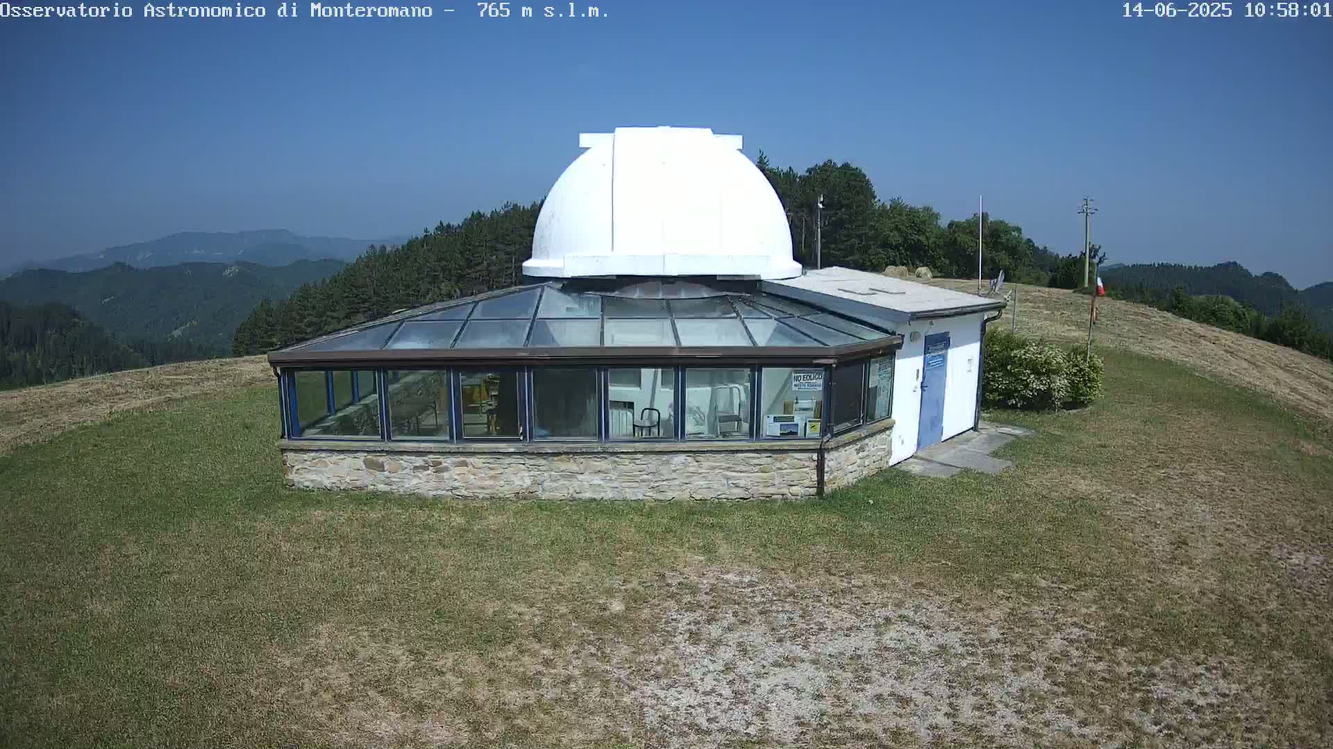 A stone building with a glass roof and a white dome sits atop a grassy hill under a clear blue sky.
