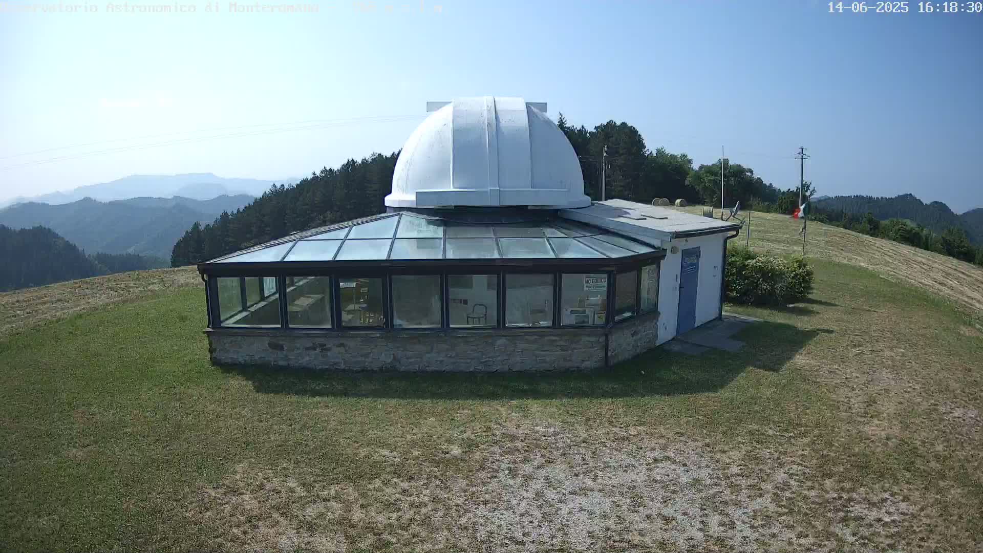 A small observatory building with a white dome sits on a grassy hilltop overlooking a range of distant mountains under a clear, sunny sky.