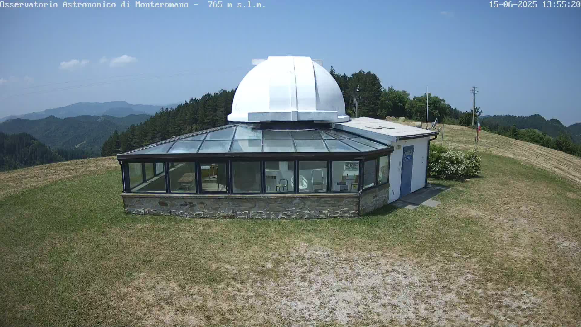 A white domed observatory building with a glass sunroom sits on a grassy hill overlooking a distant mountain range under a clear blue sky.