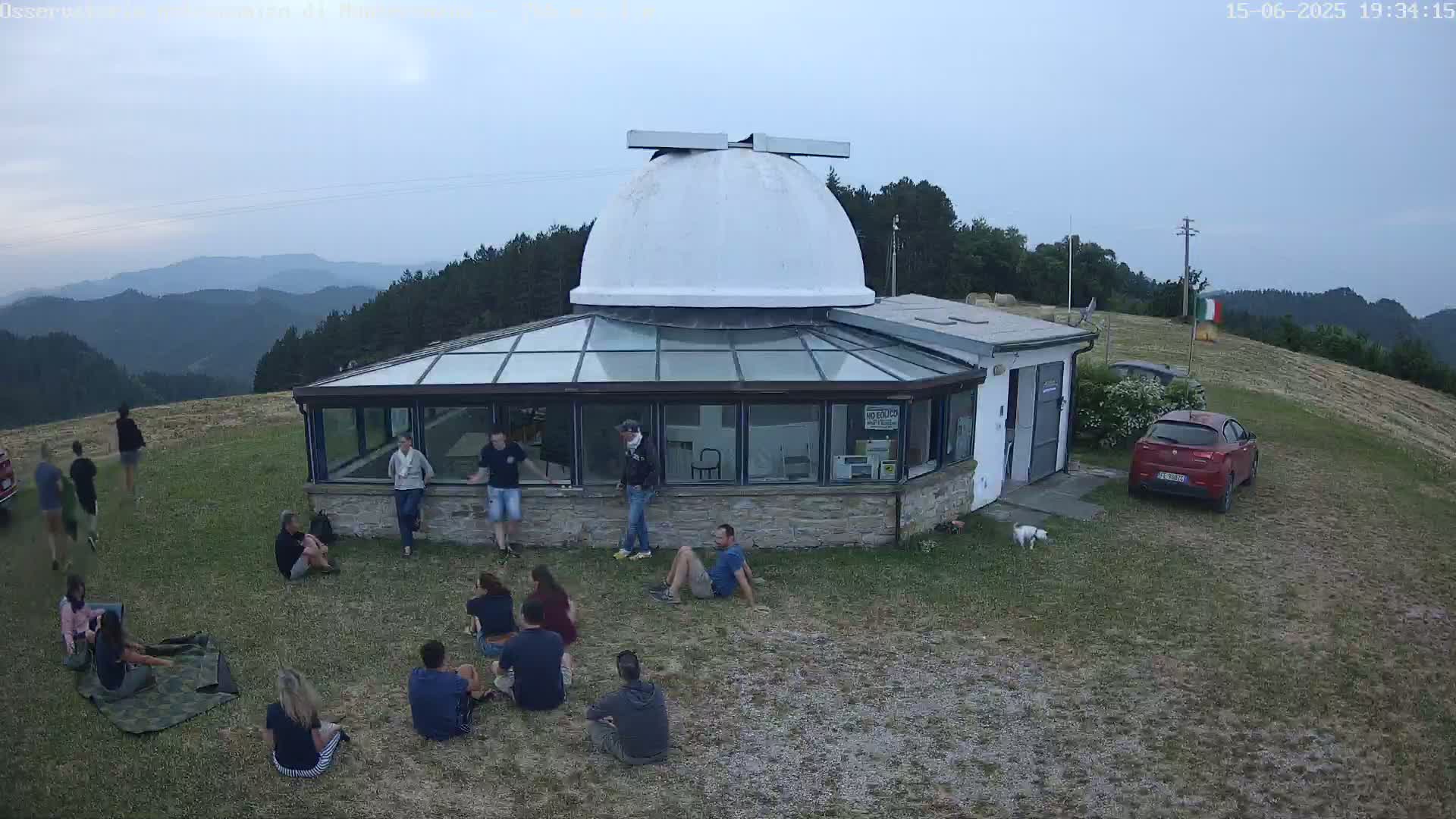A group of people are gathered outside a building with a white dome on top, overlooking a hilly landscape under an overcast sky.