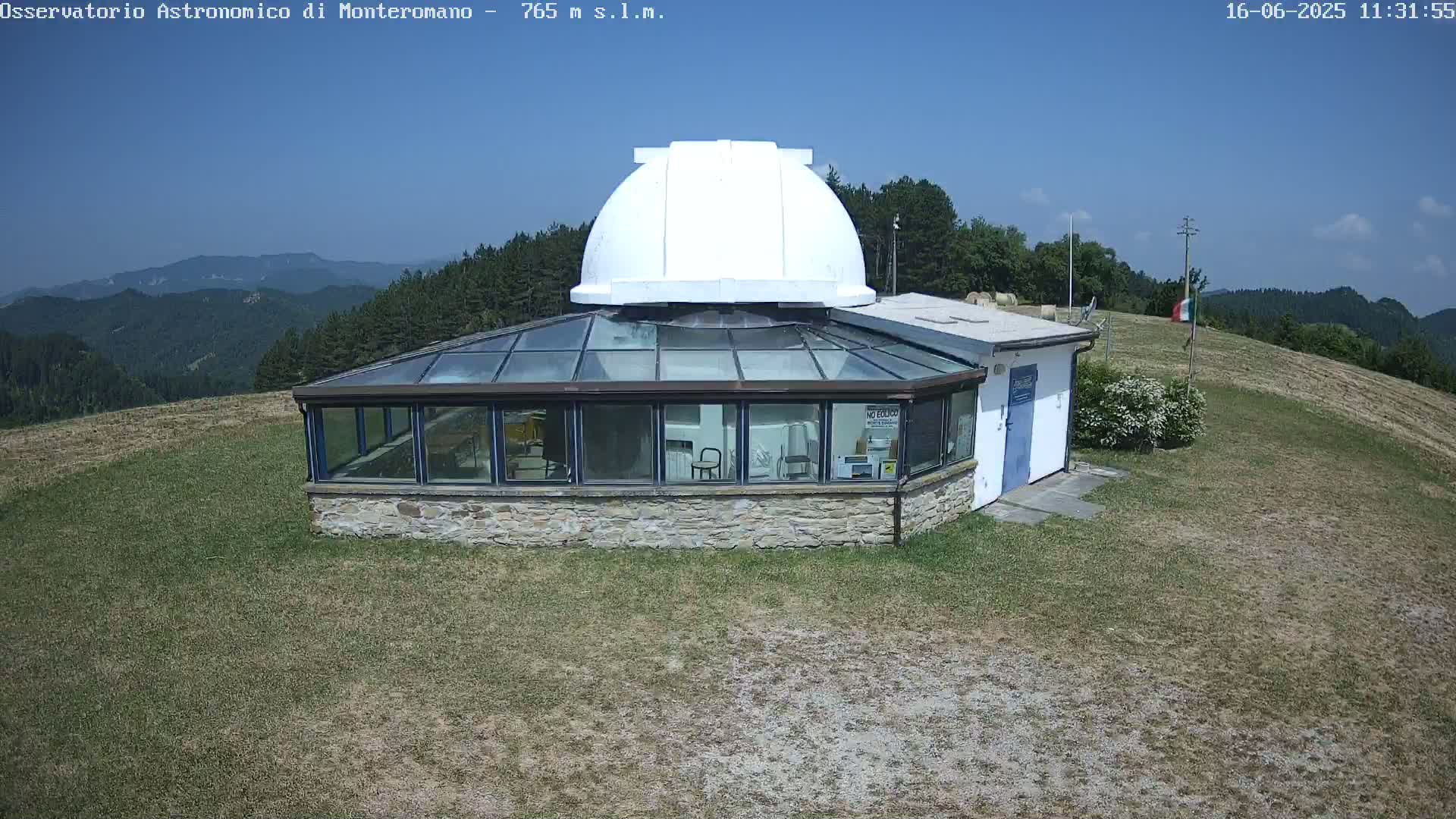 A stone observatory building with a glass enclosure and a white dome sits on a grassy hilltop under a clear blue sky.