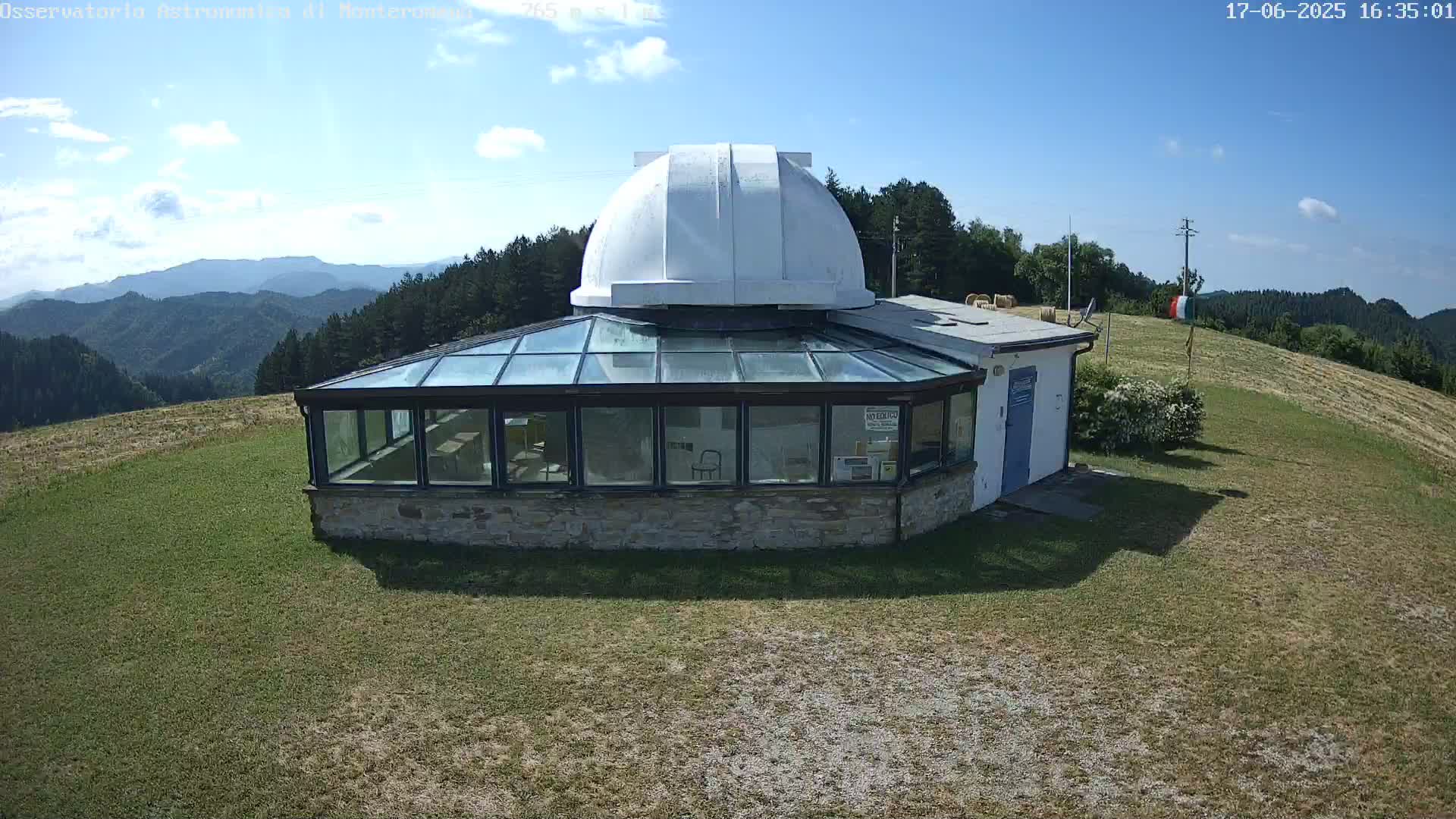 A white domed observatory building with a glass sunroom sits on a grassy hilltop under a mostly sunny sky, overlooking a distant mountain range.