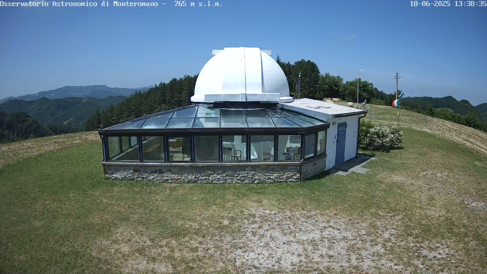 A white dome observatory with a glass-roofed addition sits on a grassy hill overlooking a mountainous landscape under a clear blue sky.