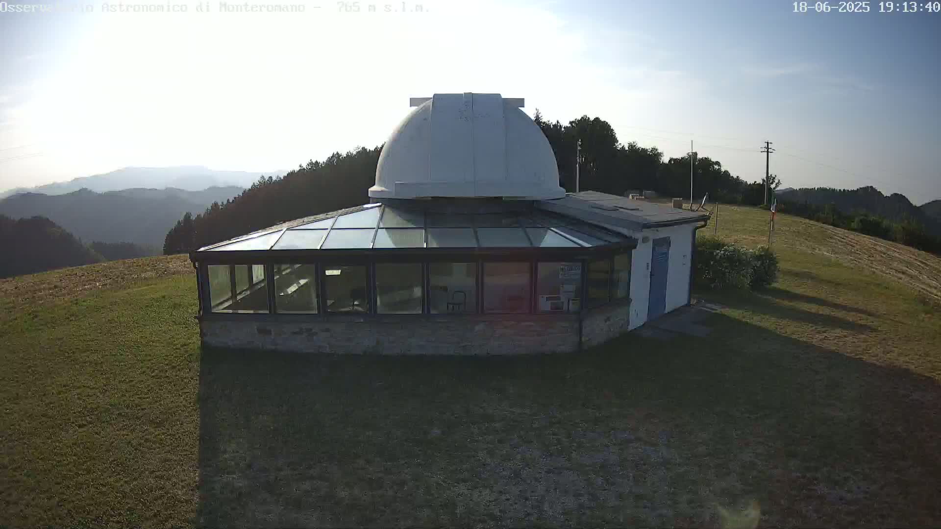 A stone building with a glass sunroom and a white dome sits on a grassy hill overlooking a hazy mountain range under a clear, sunny sky.