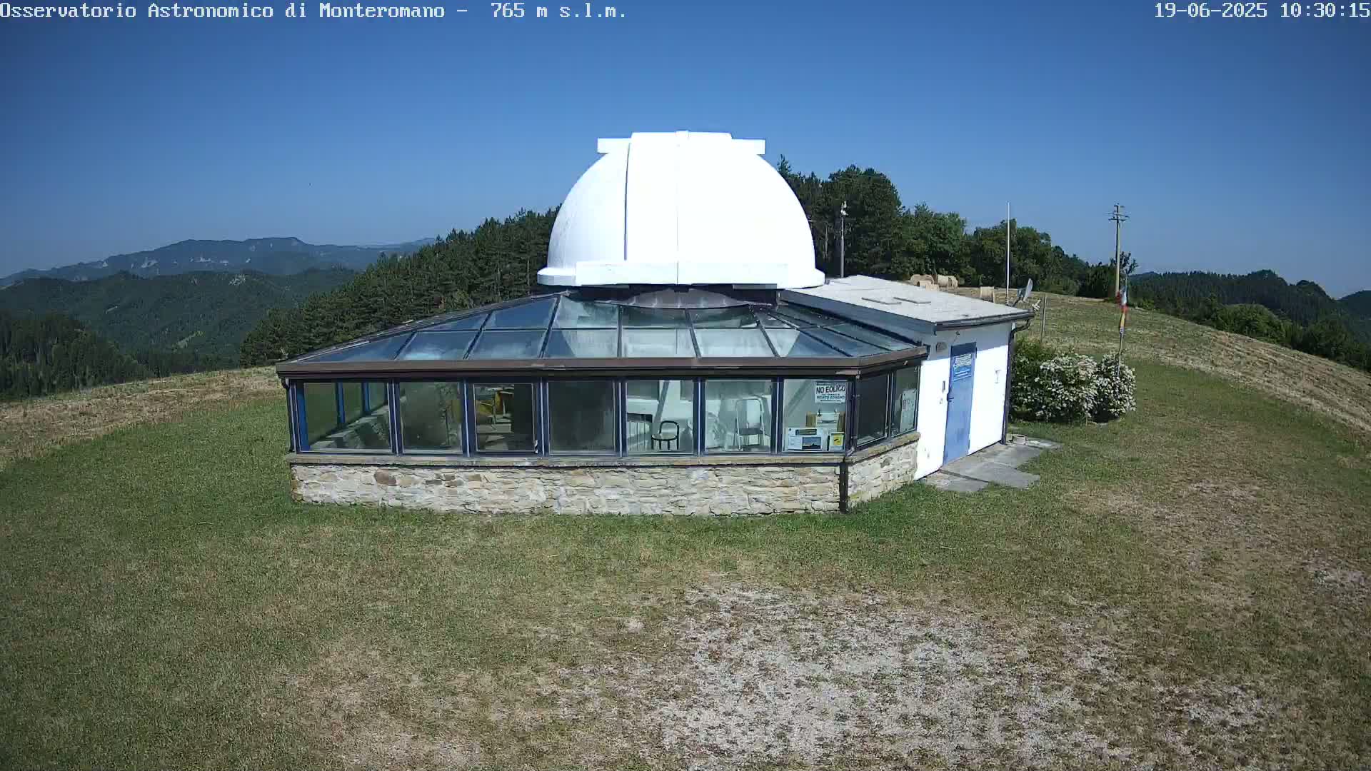 A stone building with a glass roof and a white dome sits on a grassy hilltop under a clear blue sky.