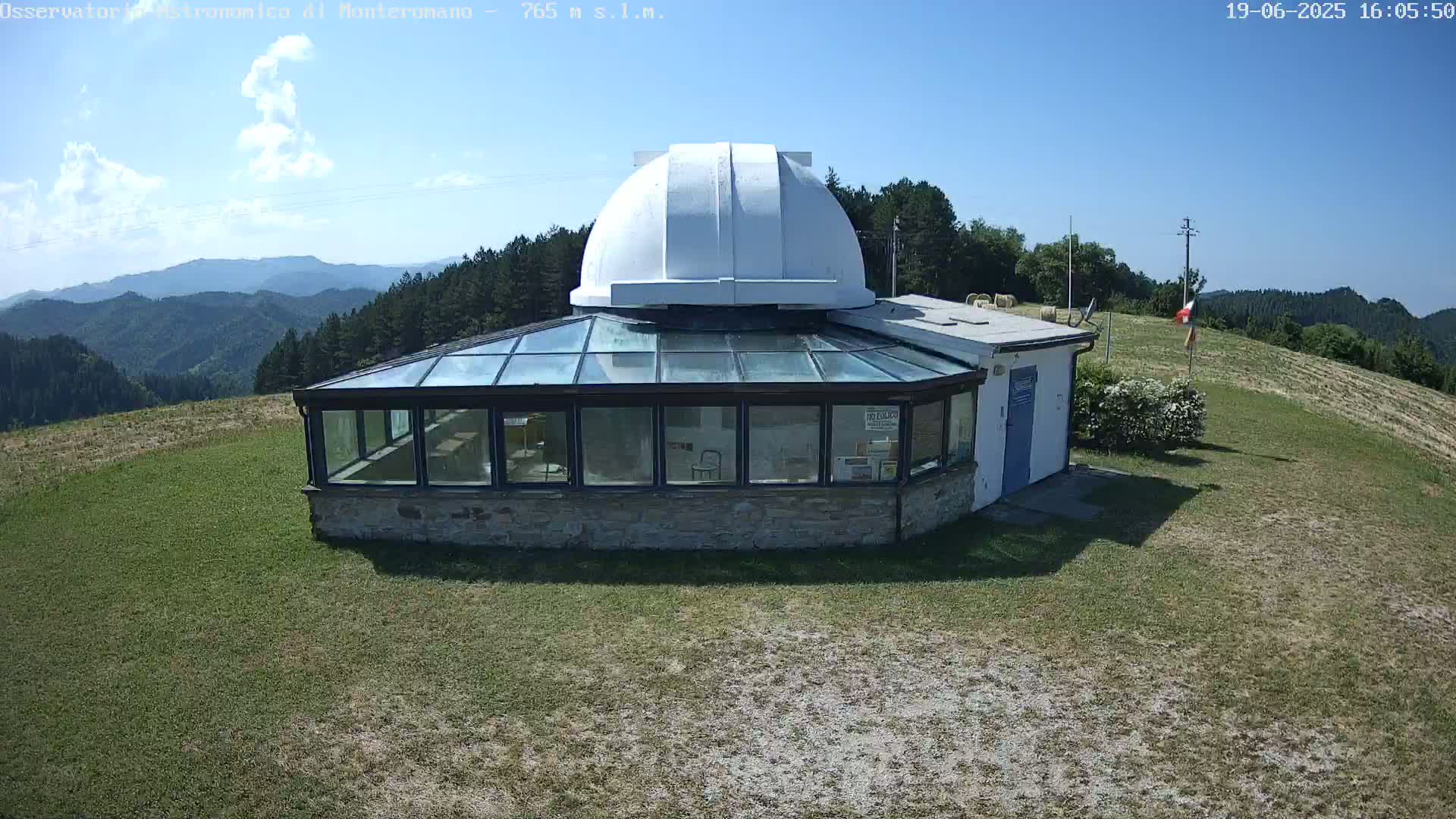 A white-domed observatory building with a glass enclosure sits on a grassy hilltop overlooking a mountain range under a mostly sunny sky.