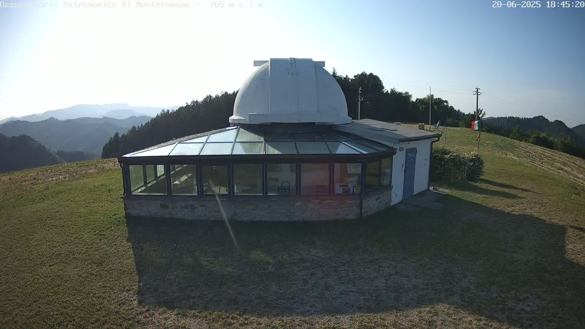 A stone building with a glass roof and a white dome atop it sits on a grassy hill overlooking a mountain range under a clear, sunny sky.