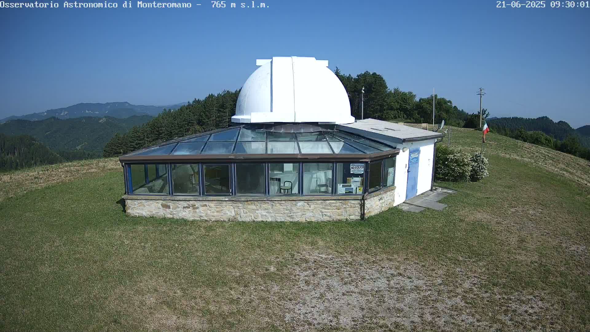A stone building with a glass enclosure and a white dome sits on a grassy hilltop under a clear blue sky.