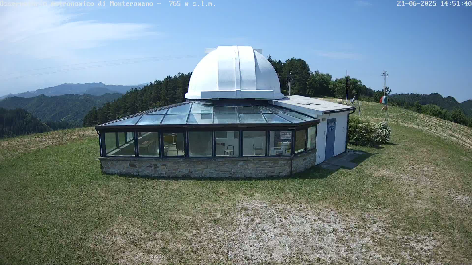 A stone building with a glass roof and a white dome sits on a grassy hilltop under a clear blue sky, overlooking a distant mountain range.