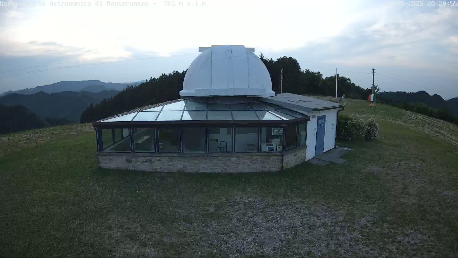A stone observatory building with a glass sunroom and a white dome sits on a grassy hilltop under a partly cloudy evening sky overlooking a mountain range.