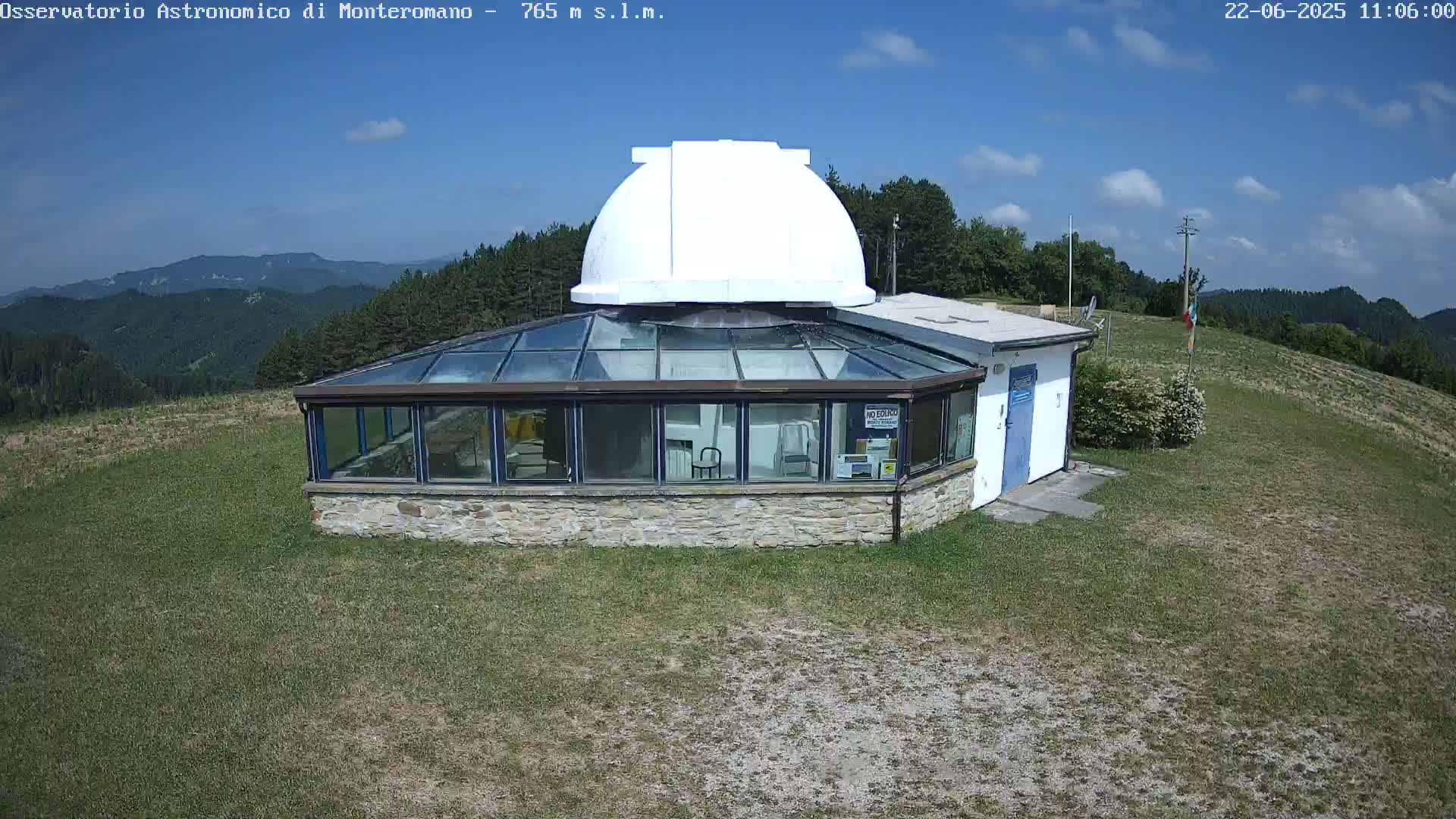 A stone building with a glass roof and a white dome on top sits on a grassy hill under a mostly sunny sky.