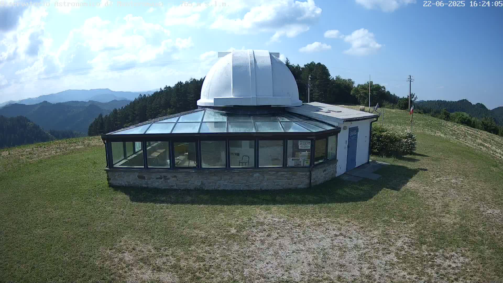 A white-domed observatory building with a glass enclosure sits on a grassy hilltop overlooking a mountain range under a partly cloudy sky.