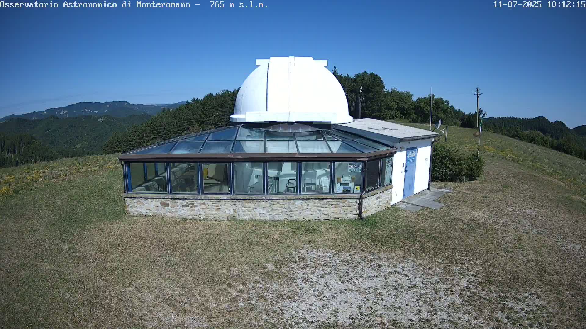 A white dome sits atop a stone building with a glass sunroom, overlooking a grassy hillside and distant mountains under a clear blue sky.