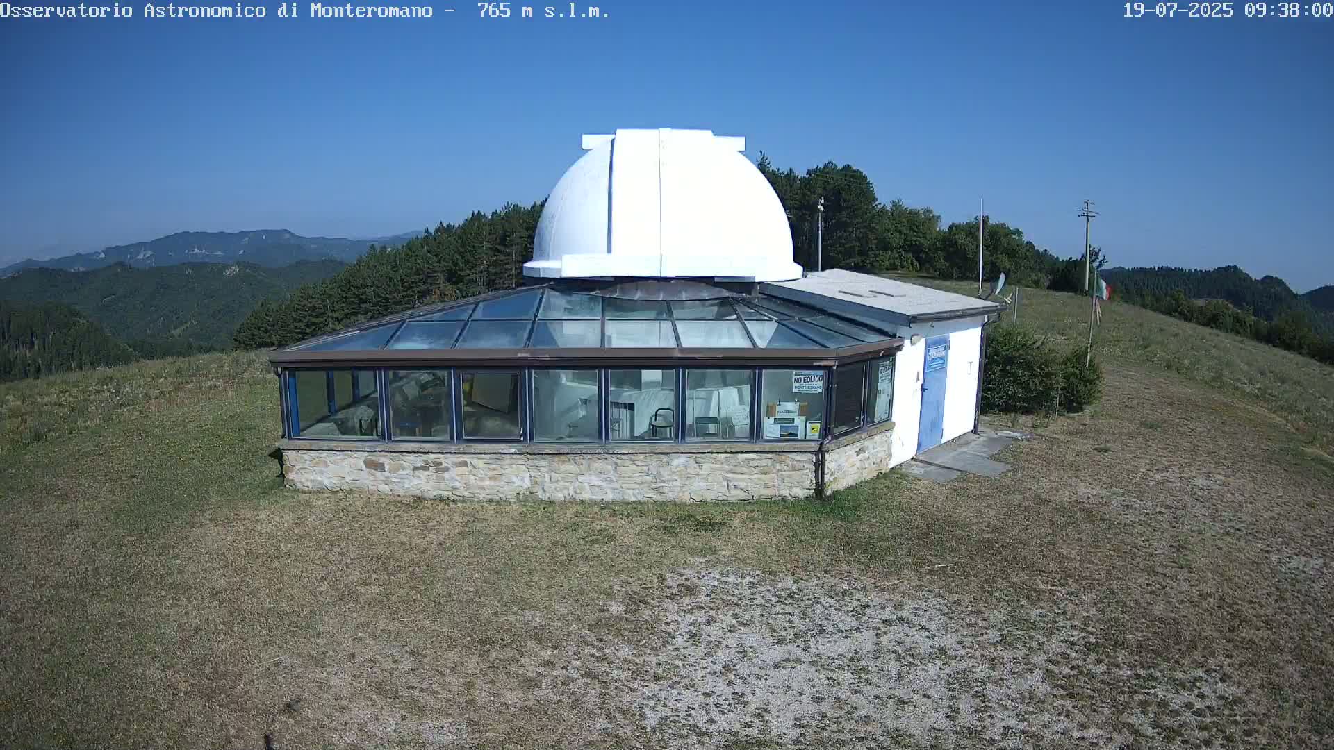 A stone building with a glass roof and a white dome sits on a grassy hilltop under a clear blue sky.