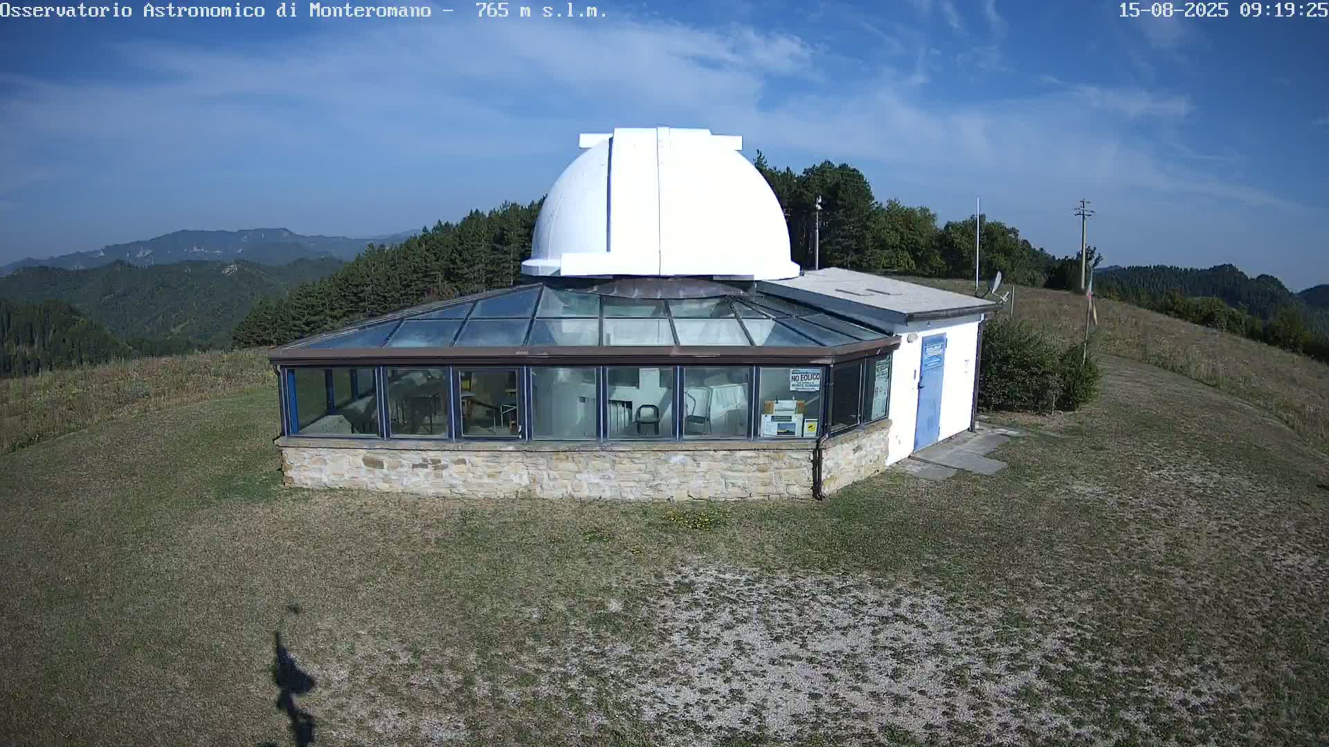 A stone building with a glass sunroom and a white observatory dome sits on a grassy hilltop under a mostly sunny sky, overlooking a mountainous landscape.