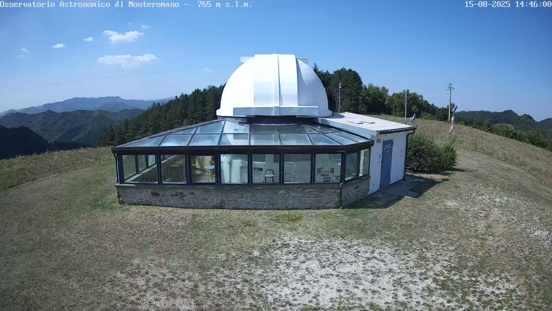 A stone building with a glass sunroom and a white observatory dome sits on a grassy hilltop under a mostly sunny sky, overlooking a mountainous landscape.