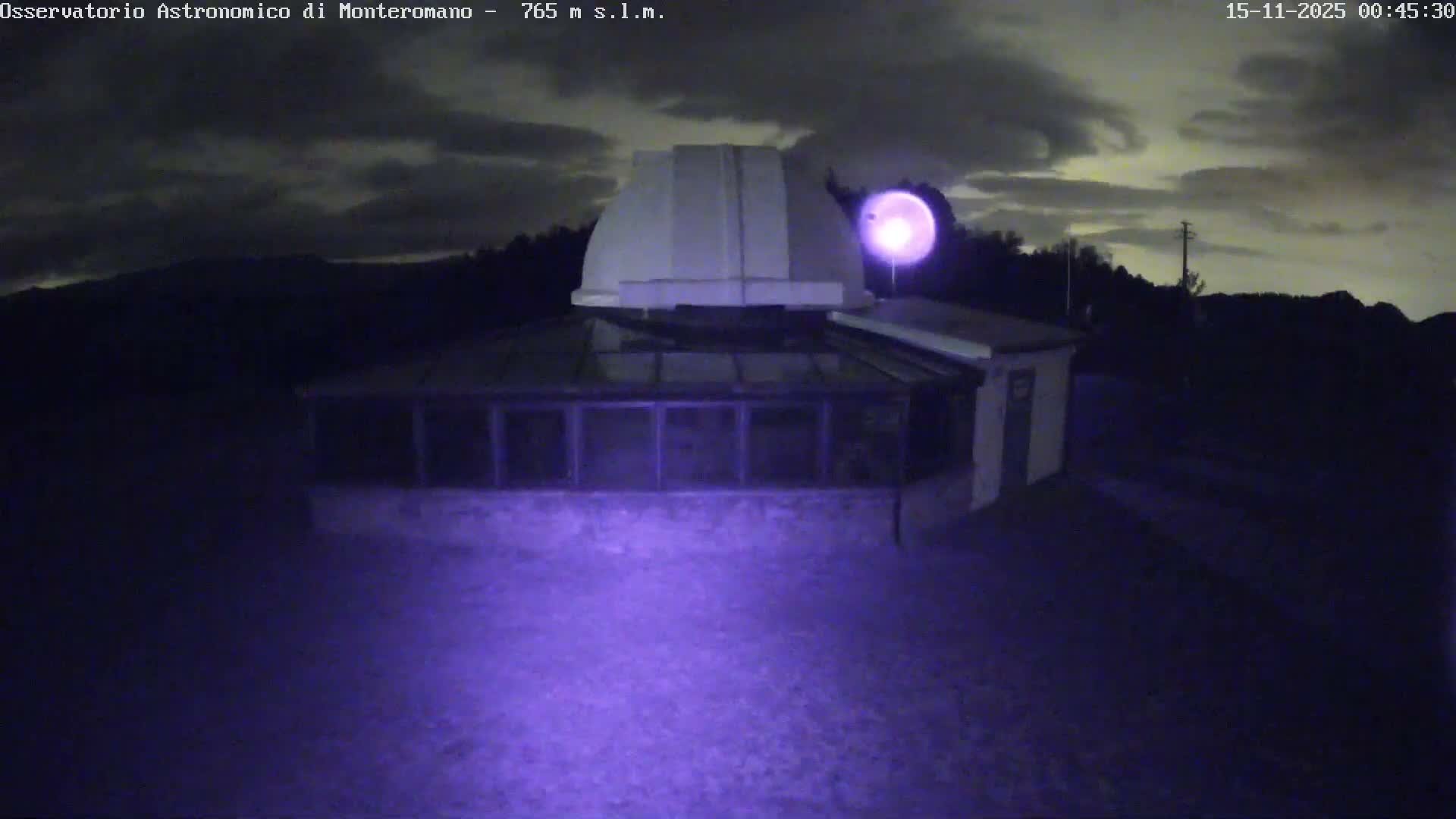 A nighttime scene captures an observatory with a prominent white dome and an adjacent structure, bathed in a soft purple glow from an orb of light, all set against a backdrop of dark hills and a cloudy sky.