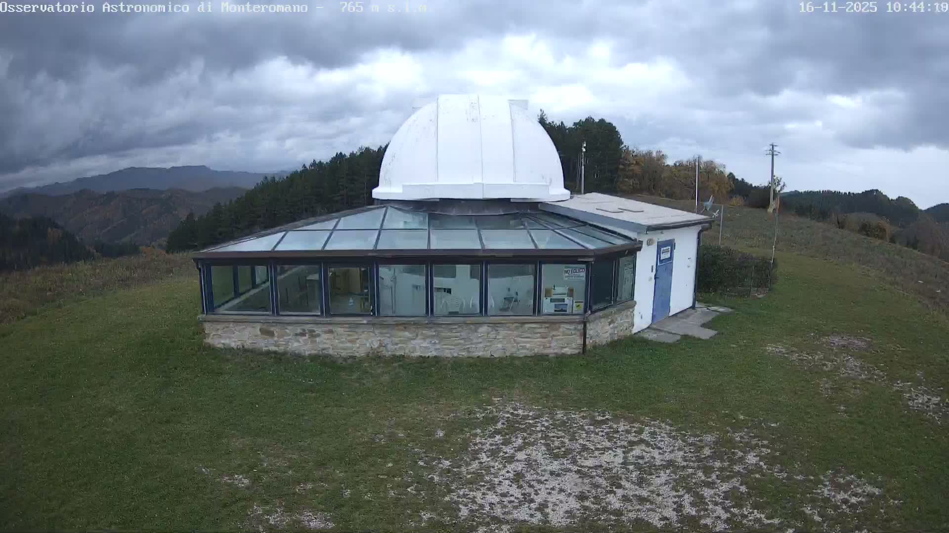 A white-domed observatory building with a glass-enclosed lower section stands on a grassy hill overlooking forested mountains under a heavily overcast, cloudy sky.