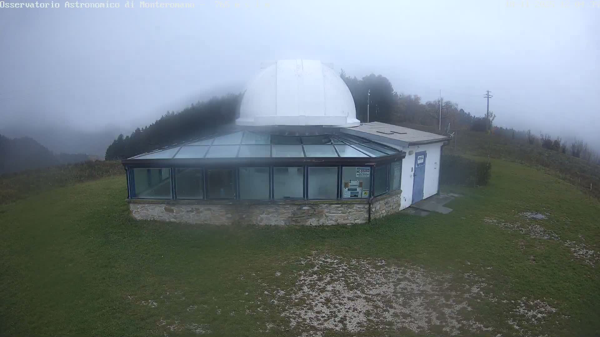 A white-domed observatory building with a glass-enclosed lower section stands on a grassy hill overlooking forested mountains under a heavily overcast, cloudy sky.