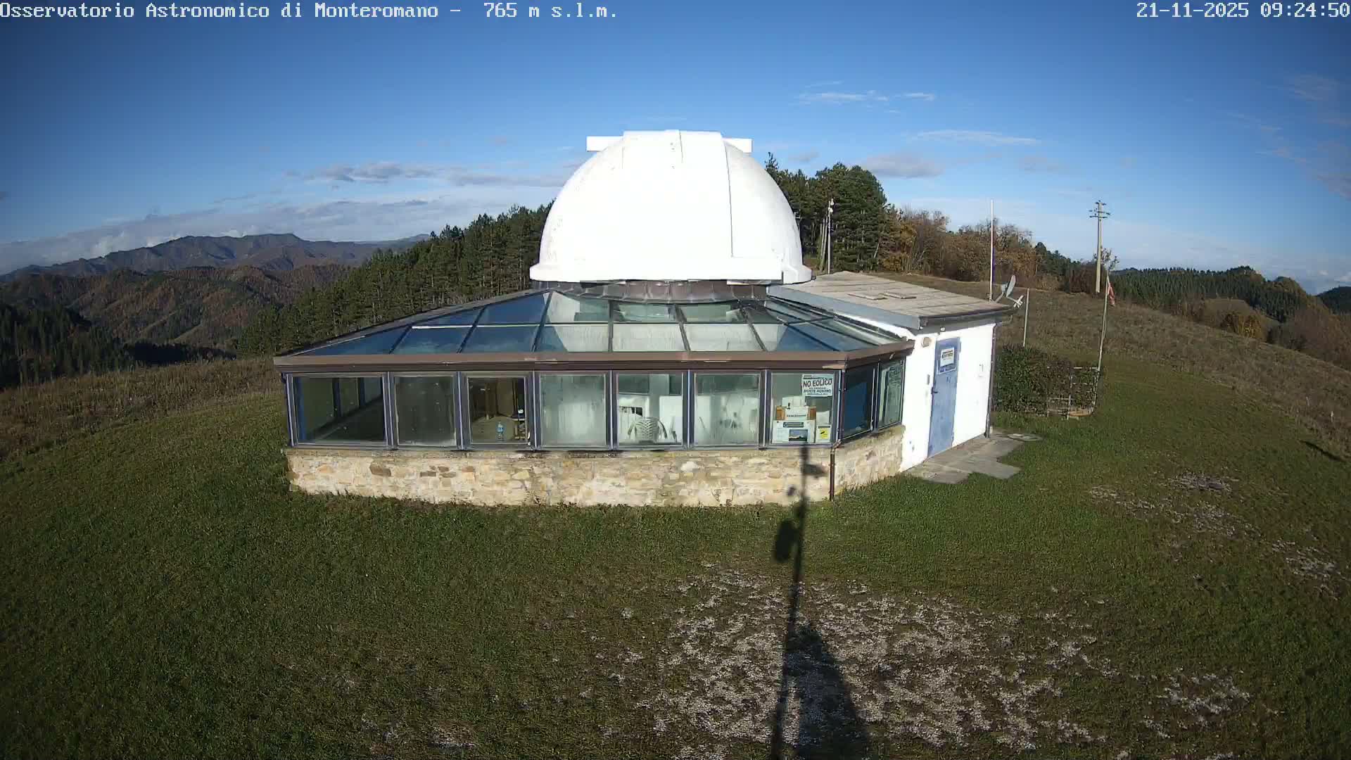 A white-domed observatory sits atop a grassy hill, surrounded by trees and distant mountains, under a clear, sunny sky with scattered white clouds.