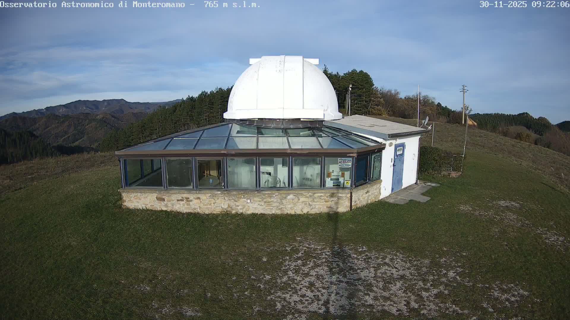 An observatory building with a white dome atop a glass-enclosed base and an attached white annex sits on a grassy hillside, overlooking tree-covered mountains under a partly cloudy sky.