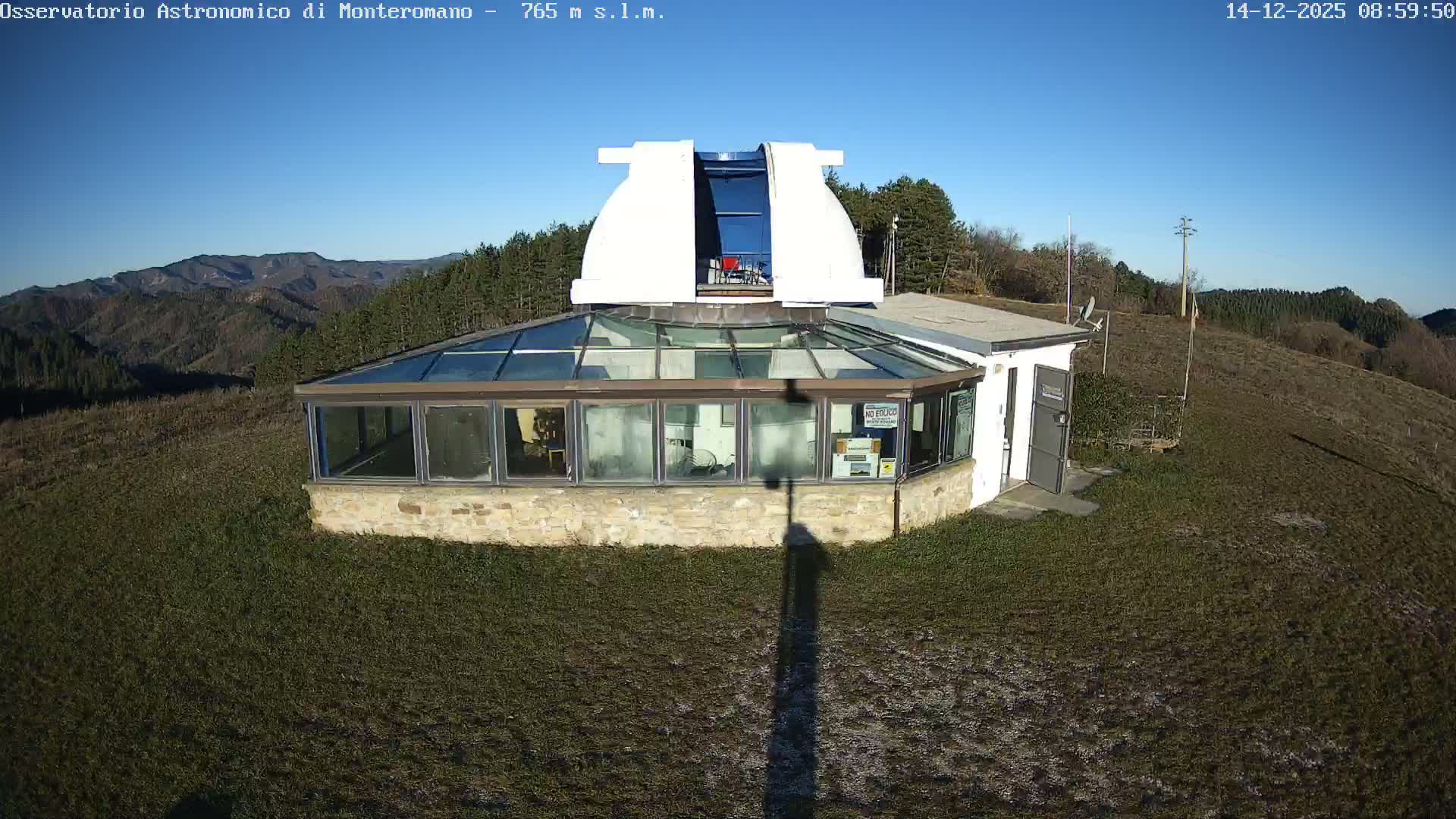 A white astronomical observatory building with an open dome and a glass-roofed section stands on a grassy hill, surrounded by a forest and distant mountains, under a clear, sunny blue sky.