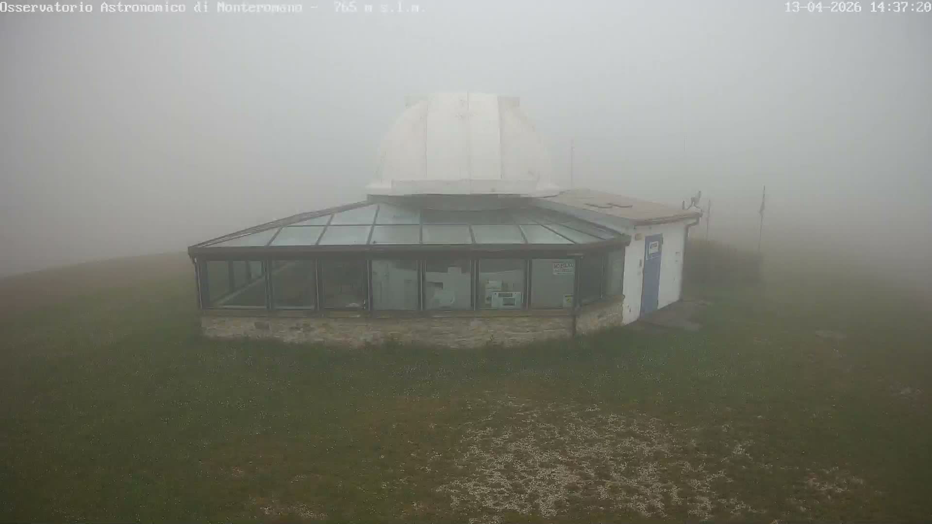 A white astronomical observatory building with an open dome and a glass-roofed section stands on a grassy hill, surrounded by a forest and distant mountains, under a clear, sunny blue sky.