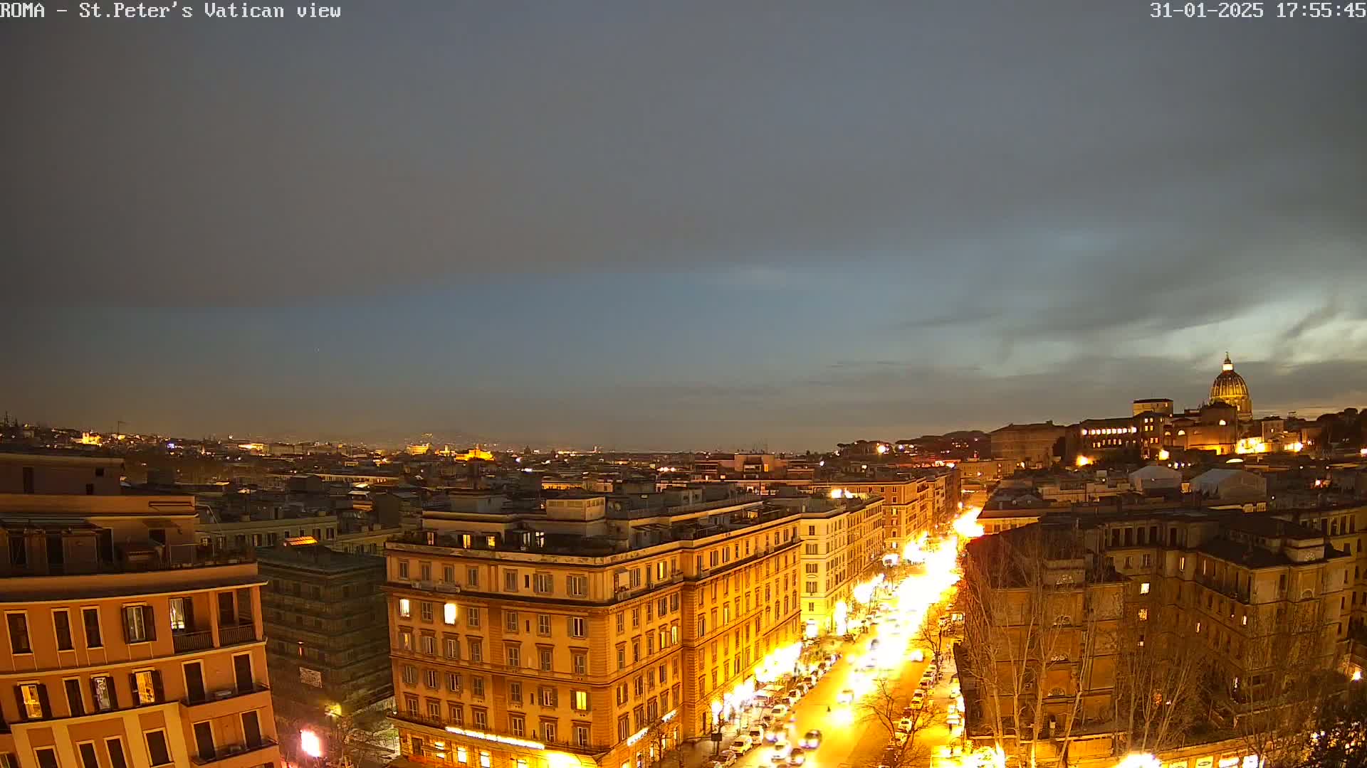 A nighttime, high-angle view of a city street with illuminated buildings and traffic under a mostly cloudy sky.